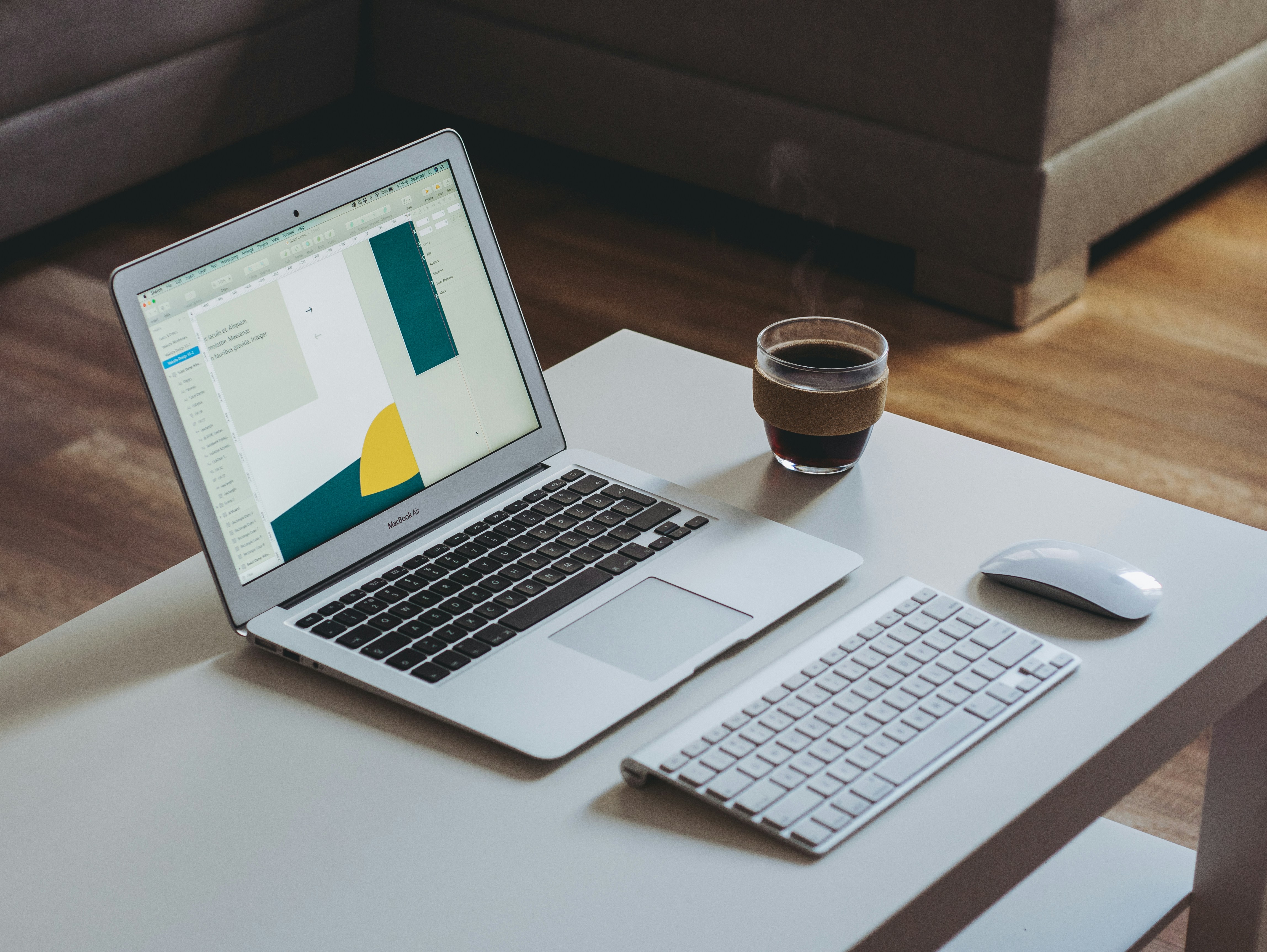 Laptop showing a design file beside a keyboard and coffee on a white desk